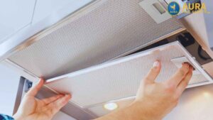 Showing the process of kitchen maintenance, a pair of hands grasp the edge of a grease filter, pulling it down from a professional-grade kitchen exhaust hood.