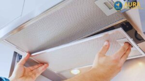Showing the process of kitchen maintenance, a pair of hands grasp the edge of a grease filter, pulling it down from a professional-grade kitchen exhaust hood.