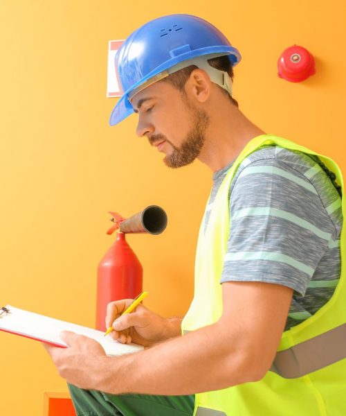 A focused technician in a blue hard hat and high-visibility vest conducting an annual fire sprinkler inspection, diligently taking notes on a clipboard beside a red fire extinguisher, against a vibrant orange wall.