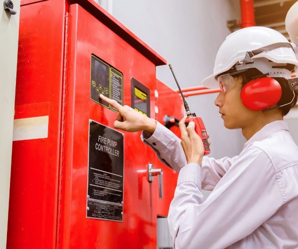 A professional fire safety engineer conducting a fire sprinkler system inspection at a control panel, wearing a white hard hat, earmuffs, and safety glasses, with red fire suppression systems in the background.